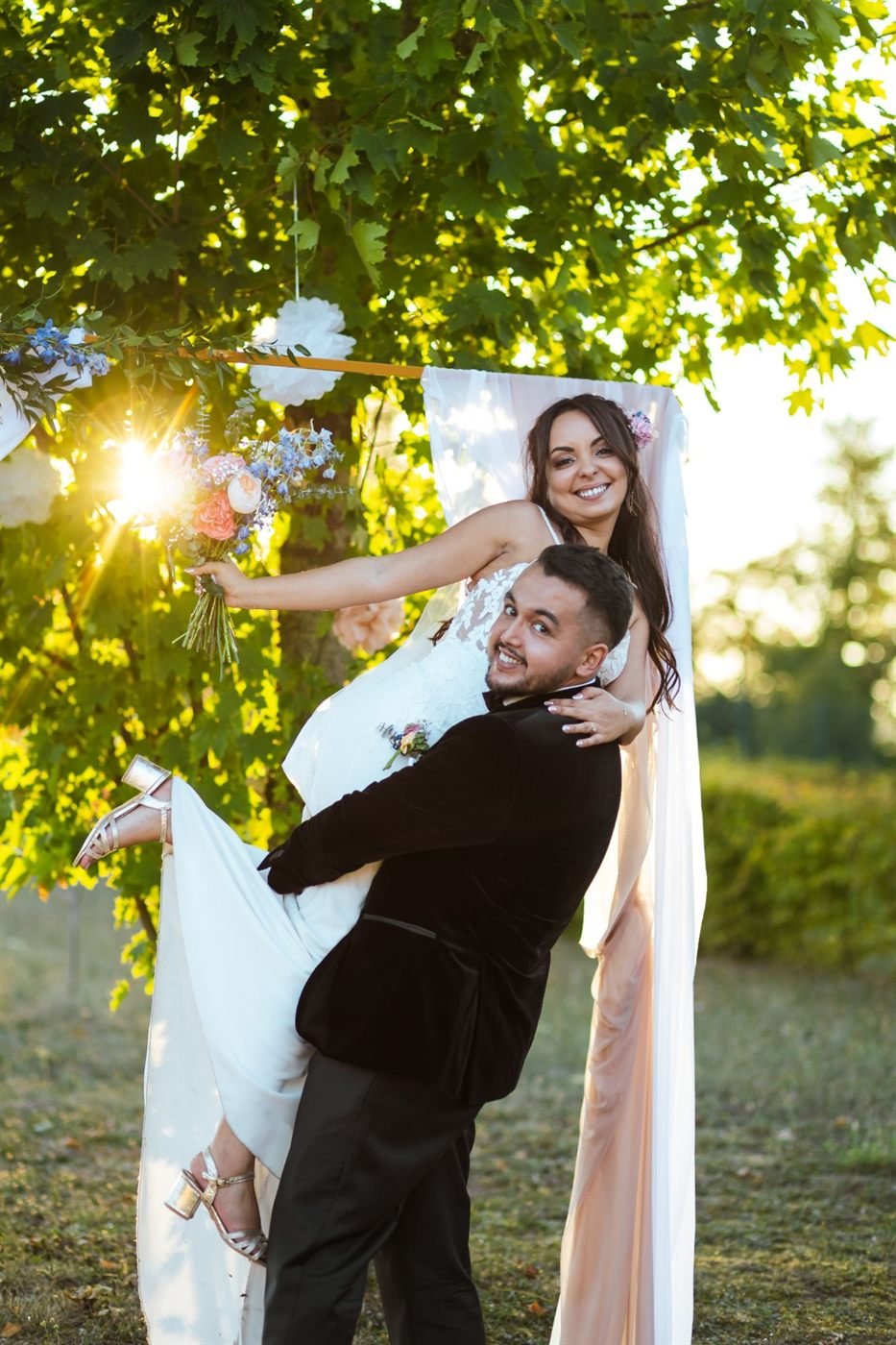 Le marié porte la mariée en riant au coucher du soleil le jour du mariage
