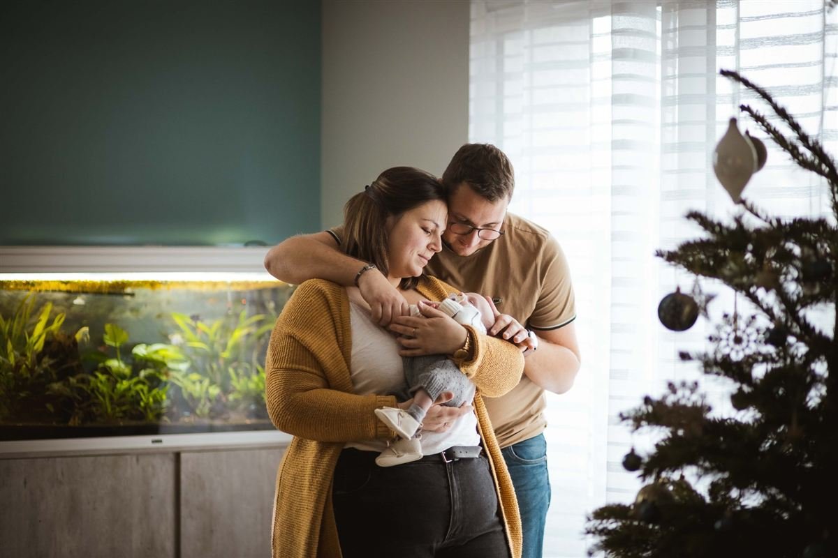Séance naissance à domicile : des parents enlacés tiennent leur nouveau-né près du sapin de Noël
